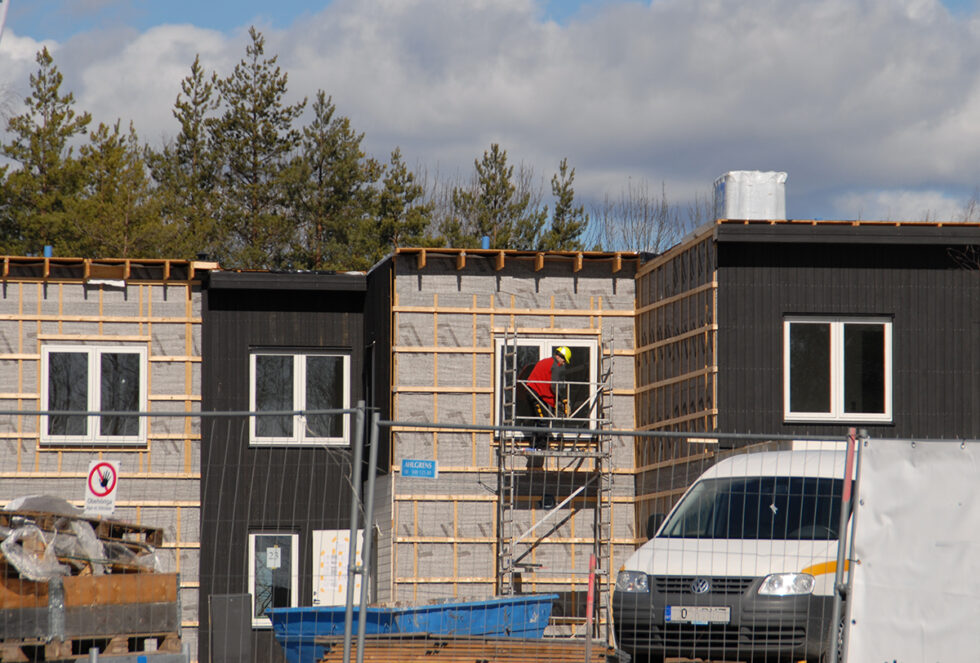 Byggarbetsplats där byggandet av tvåvåningshus pågår. Man ser en byggnadsarbetare i hjälm och arbetskläder på en ställning.