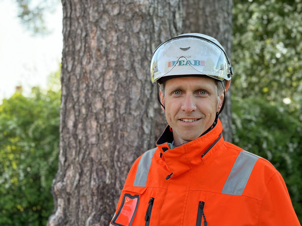 Porträtt tagit utomhus av en man i orange varseljacka och vit bygghjälm. Peab tryckt på hjälmen.