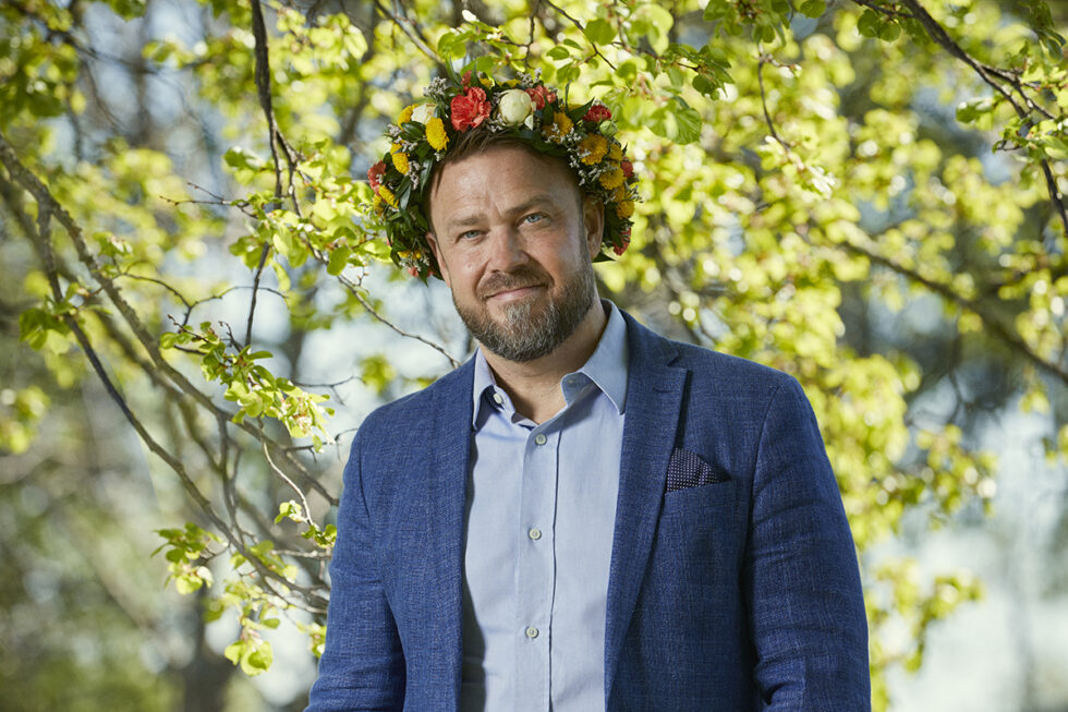 Kim Söderström klädd i kavaj och med blomsterkrans på huvudet står framför grönskande växter