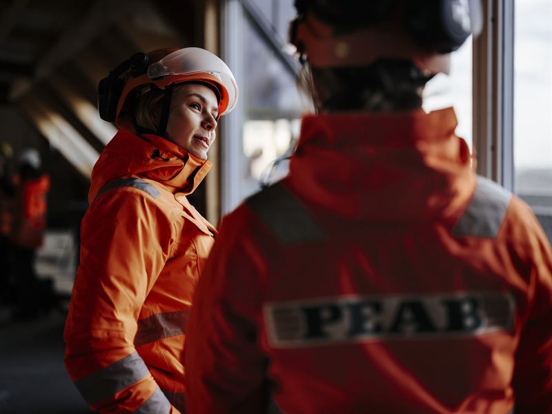 Två peabmedarbetare i bygghjälpar och orangea jackar loggan på ryggen.